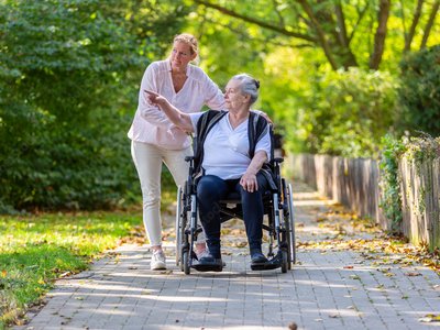 Frau macht einen Spaziergang mit einer älteren Dame im Rollstuhl.