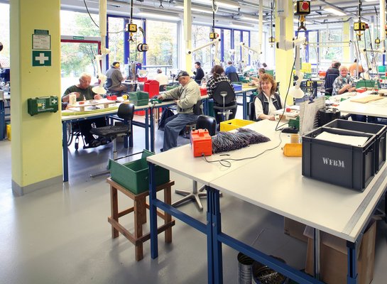 Die Werkstatt - An den Werkbänken sitzen die Mitarbeiter und arbeiten.