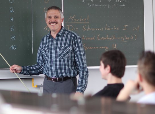 Ausbilder erklärt etwas an der Tafel und erklärt lächelt Schüler an.
